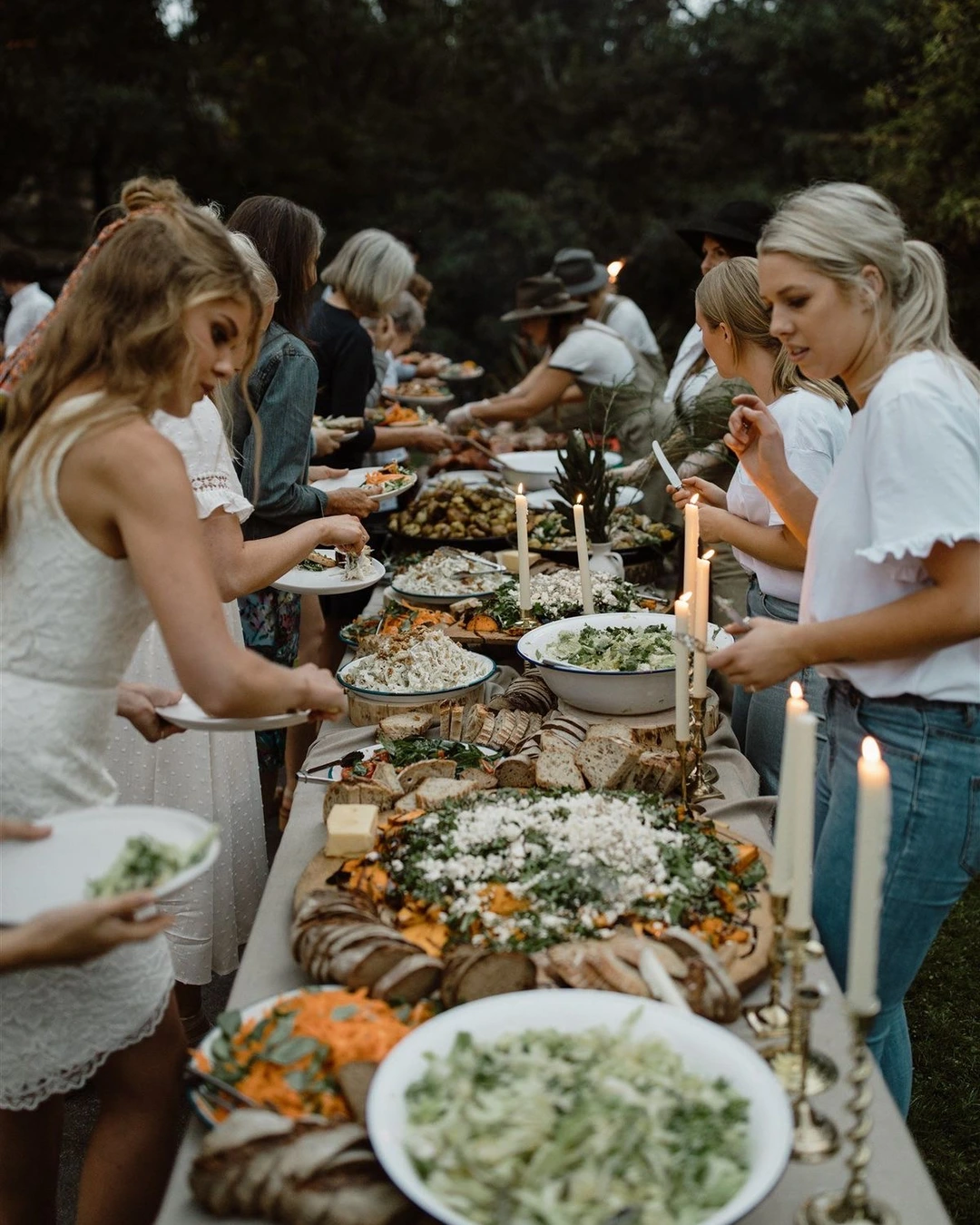 Festgesellschaft an einer langen Tafel im Grünen mit Kerzen und weißen Blüten